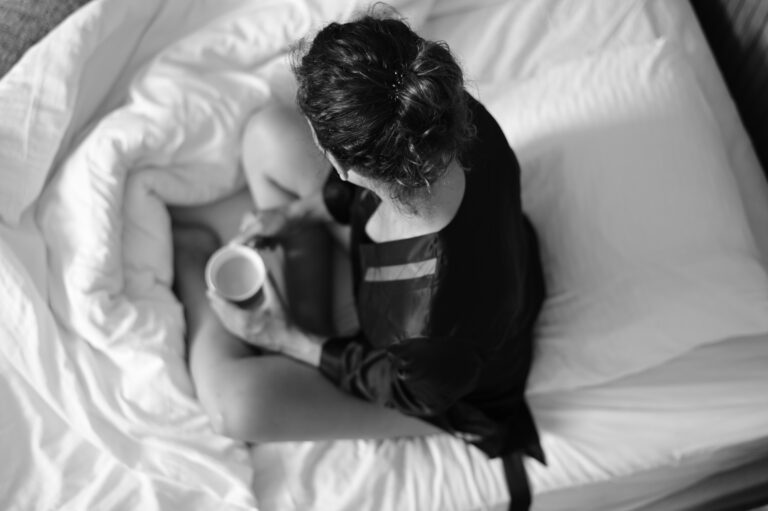 Kvinne med kaffekopp sitter i seng, ovenfra i svart-hvitt